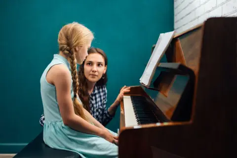Dziewczynka z warkoczem uczy się gry na pianinie pod okiem nauczycielki. Nuty na pulpicie wskazują na lekcję muzyki.