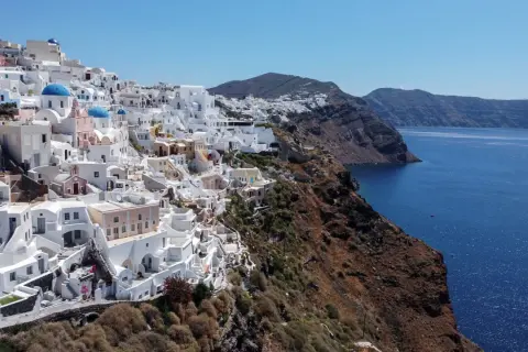 Białe domy Santorini na klifie, z błękitnymi kopułami kościołów, nad lazurowym morzem. Krajobraz jak po trzęsieniu ziemi, ale pełen życia.