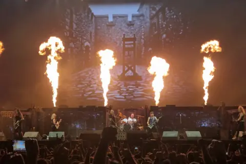 Płomienie buchają na scenie, gdzie grają zespoły rockowe. W tle widać zamek i gilotynę.