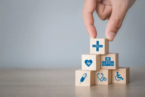 Dłoń układa kostki symbolizujące opiekę zdrowotną. Może to oznaczać wsparcie dla osób z lekkim stopniem niepełnosprawności, np. zasiłek z MOPS.