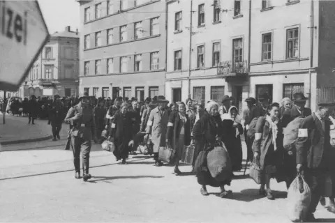 Människor med armbindlar ledsas förbi Schindlers fabrik. En soldat går framför.