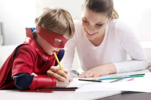 Chłopiec w stroju superbohatera rysuje, a mama z uśmiechem wspiera go, pokazując, jak zachęcić dziecko do nauki przez zabawę.