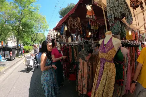 Persone curiosano tra le bancarelle colorate della fiera di San Ciriaco, ammirando abiti etnici e tessuti vivaci sotto il sole.