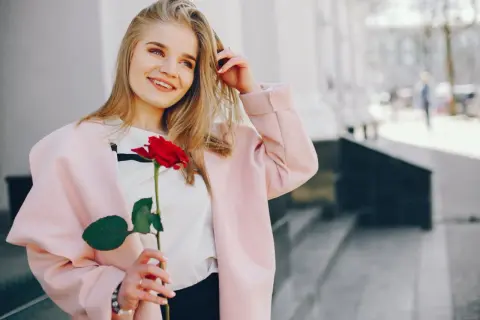 Uśmiechnięta blondynka w różowym płaszczu trzyma czerwoną różę. Idealna stylizacja, jak się ubrać na randkę.