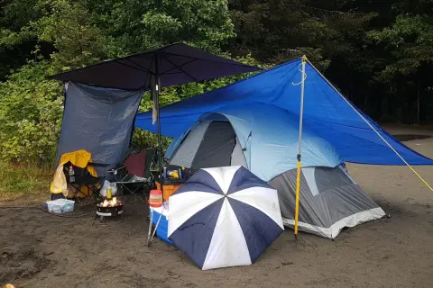 Campingplats med tält, eldstad och paraply. Ved torkar under presenning.