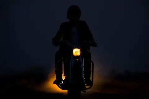 Silhouette d'une personne conduisant une moto dans l'obscurité, le phare jaune illuminant la route. L'aventure de conduire la nuit.
