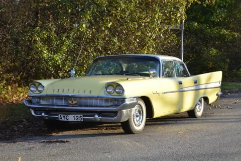 Gul Chrysler Saratoga från 1950-talet, med kromdetaljer och vita däcksidor, parkerad vid en lummig buske.