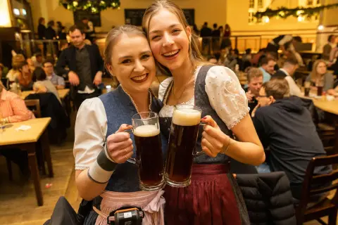 Zwei fröhliche Frauen in Tracht halten je ein großes, gestauchtes Bier. Im Hintergrund ist eine belebte Gaststube zu sehen.