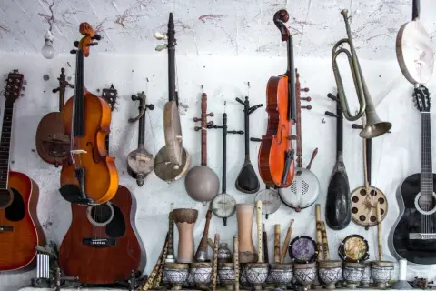 Różnorodne instrumenty strunowe, w tym gitary, skrzypce i tradycyjne lutnie, wiszą na ścianie obok bębnów i fletów.