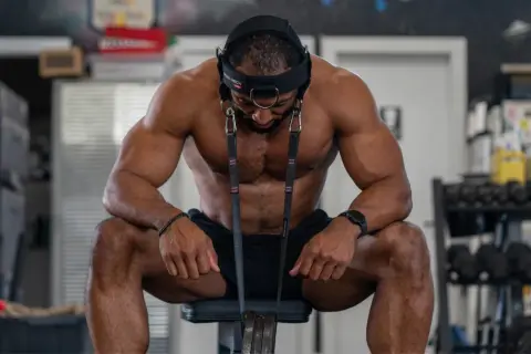 A muscular man sits, head harness on, preparing for neck exercises. He looks down, focused on his next move.