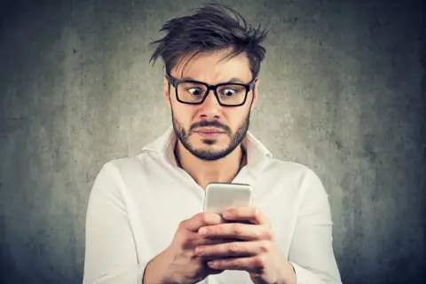 Przerażony mężczyzna w okularach czyta na telefonie o tym, co grozi za oczernianie drugiej osoby.