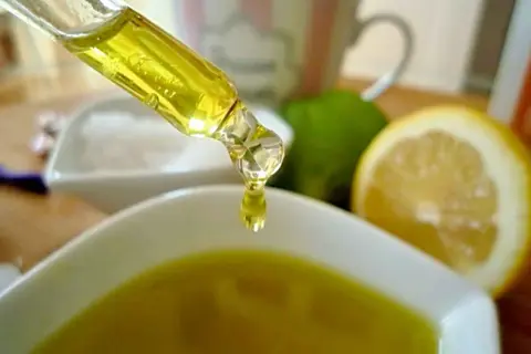Tropfen eines goldenen Öls fallen aus einer Pipette in eine Schale. Zitronen und Limetten im Hintergrund deuten auf natürliche Inhaltsstoffe für Kosmetik hin, wie z.B. Zitronensäure.