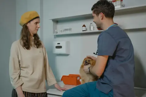 Kobieta z psem u weterynarza. Lekarz bada małego, puszystego pieska, a właścicielka obserwuje.