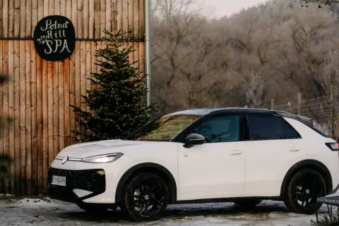 Biały, mały SUV zaparkowany obok drewnianej chaty z napisem "Polna Hill SPA" i choinką ozdobioną lampkami.