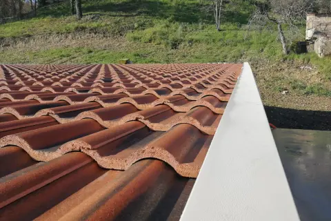 Tegole per tetti a bassa pendenza, color terracotta, disposte in file ordinate su un tetto. Sullo sfondo, un pendio erboso.
