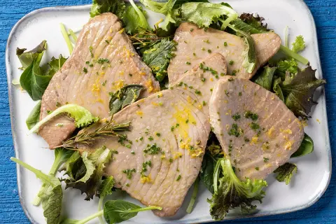 Fette di tonno al forno, dorate e succulente, servite su un letto di insalata fresca con scorza di limone e erbe aromatiche.