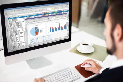 Mężczyzna pracuje przy komputerze, analizując dane w arkuszu kalkulacyjnym, przypominającym ten z programu **jeżeli excel**. Na ekranie widoczne są wykresy i tabele.