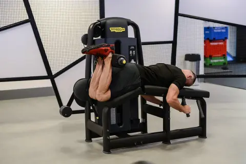 Mężczyzna wykonuje ćwiczenie na maszynie do uginania nóg leżąc (hamstring curl), budując siłę tylnej części uda.