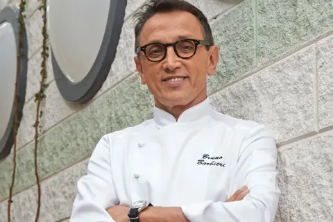 Bruno Barbieri, chef stellato Michelin, sorride con le braccia conserte davanti a un muro di pietra.