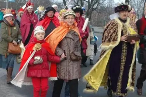Kolorowe stroje Trzech Króli na ulicznym orszaku. Dziecko w czerwonym płaszczu i koronie, obok dorosłe osoby w bogatych szatach.