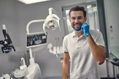 Uśmiechnięty dentysta w gabinecie, gotowy do pracy. Zastanawiasz się, ile trwa plombowanie zęba?