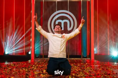 Vincitore del finale MasterChef 2026 in ginocchio tra coriandoli dorati, con le braccia alzate e il logo di MasterChef sullo sfondo.