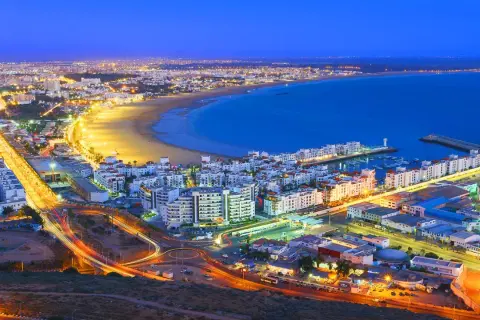 Agadir, Marocko, upplyst vid skymningen. En livlig stad vid havet med en strandpromenad, en marina och ljusspår från trafiken.
