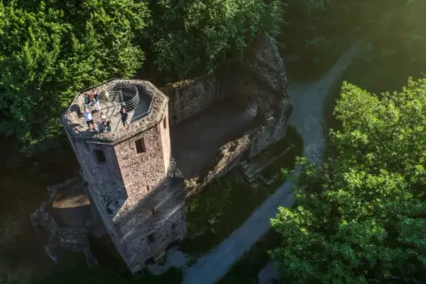 Blick von oben auf die Ruine der Barbara Kapelle, umgeben von grünen Bäumen. Eine Gruppe von Menschen genießt die Aussicht vom Turm.