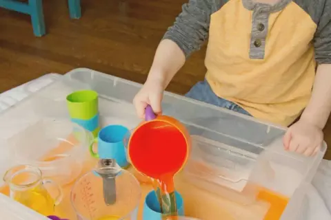 A child enjoys Montessori water play, pouring orange liquid from a colorful pitcher into a blue cup.