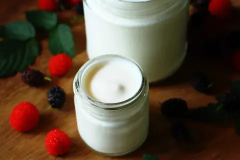 Dos frascos de yogurt casero cremoso, decorados con moras y hojas verdes. Una guía visual para aprender como hacer yogurt casero.