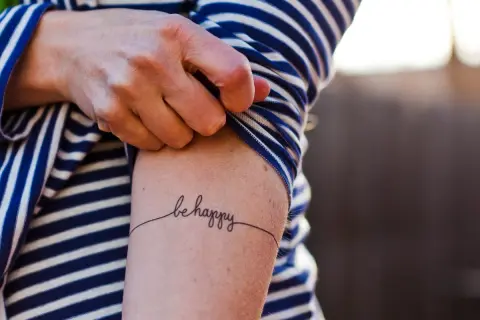 Un brazo con una camiseta a rayas azules y blancas muestra un delicado tatuaje de caligrafía que dice "be happy", una forma de arte para transformar cicatrices.