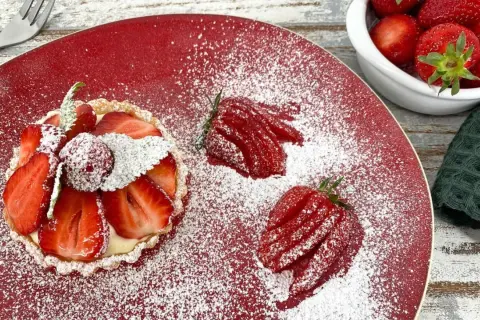 Zuckersüße Erdbeer Tartelettes, bestäubt mit Puderzucker, auf rotem Teller. Daneben frische Erdbeeren und eine kleine Schale voller roter Früchte.