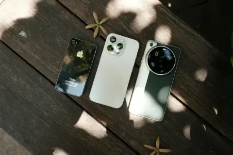 Trzy telefony do zdjęć: stary iPhone, nowy iPhone i smartfon z aparatem Leica, leżące na drewnianym stole.