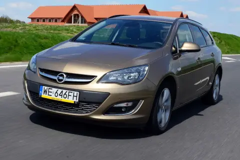 Brązowy Opel Astra J automat w wersji kombi jedzie po drodze. W tle widać domy z czerwonymi dachami.