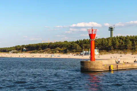 Mrzeżyno atrakcje: plaża, morze i charakterystyczna czerwona wieża nawigacyjna na falochronie. W tle las i zielona wieża.