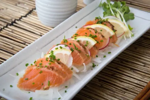 Tataki de salmón fresco, finamente cortado y adornado con rodajas de limón, rábano y cebollino, servido sobre una cama de finas hebras de cebolla.