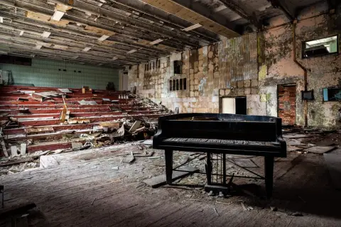 Un piano noir trône au milieu d'une salle délabrée, vestiges de maisons abandonnées. Les sièges rouges sont brisés, le sol jonché de débris.