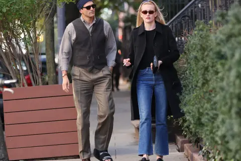 Coppia passeggia, lui con gilet e fascia, lei con cappotto e jeans a zampa. Un look che ricorda lo stile di Calvin Klein stilista.