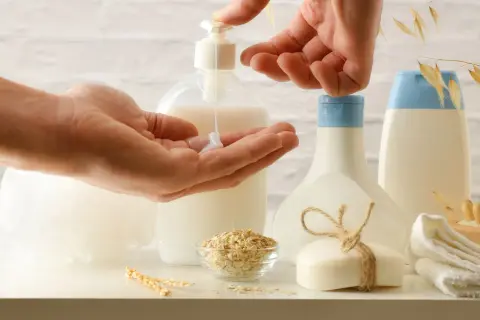 Hands dispensing lotion from a pump bottle into a palm, with oat flakes, soap, and bottles in the background.