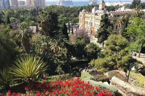 Vista panorámica de un jardín exuberante con flores rojas y palmeras, un edificio histórico y el mar al fondo en Málaga.