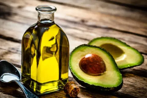 Botella de aceite de aguacate para la piel, junto a un aguacate cortado y una cuchara con aceite.