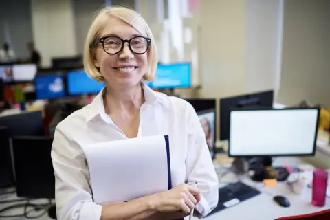 Uśmiechnięta kobieta z blond włosami i okularami, trzymająca teczkę. Jej emerytura z Anglii w Polsce przebiega pomyślnie.