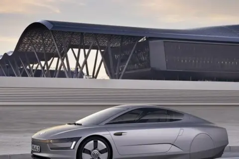 Srebrny, futurystyczny vw xl1 z charakterystycznym dachem i nowoczesnym budynkiem w tle.