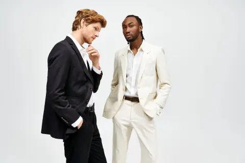 Due uomini in abiti eleganti. Uno con giacca nera e pantaloni neri, l'altro con giacca e pantaloni color crema. Il **regular fit significato** è un taglio classico e confortevole.