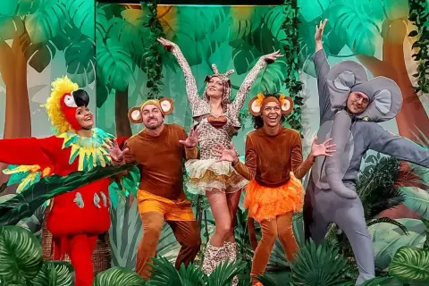 Zuzanna w stroju żyrafy i jej przyjaciele – papuga, małpki i słoń – bawią się na scenie podczas wesołego spektaklu.