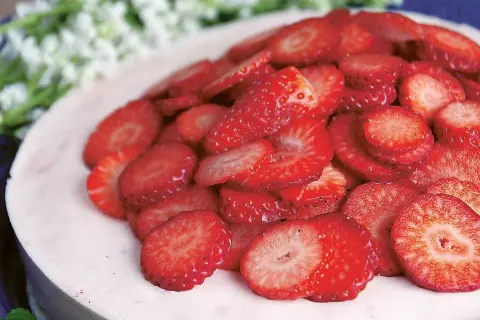 En läcker jordgubbsmousse tårta, toppad med färska jordgubbsskivor och vita blommor i bakgrunden.