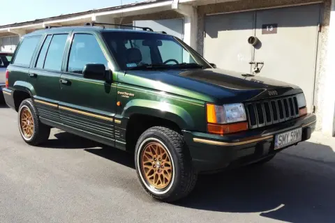 Zielony Jeep Grand Cherokee Limited z lat 90. na złotych felgach, zaparkowany przed garażami.