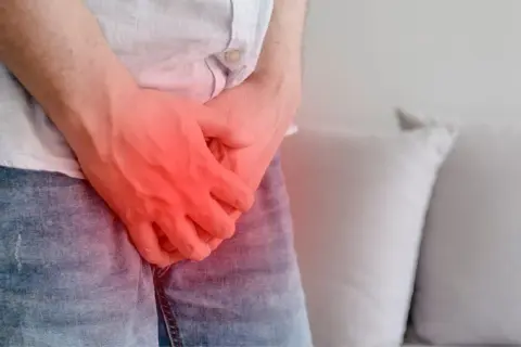 Mężczyzna w dżinsach i koszuli, z rękami na kroczu, gdzie widoczny jest czerwony obszar bólu. Może to symbolizować nietrzymanie moczu u mężczyzn po alkoholu.