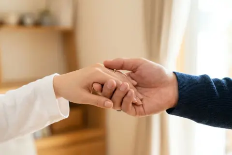 Zwei Hände halten sich fest, ein Ring am Finger. Die Nähe ist tröstlich, doch die Angst vor einer Fernbeziehung schwingt mit.