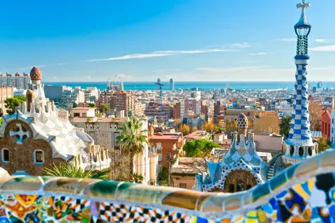 Widok na kolorowe budowle Parku Güell i panoramę miasta, gdzie leży Barcelona, z morzem w tle.